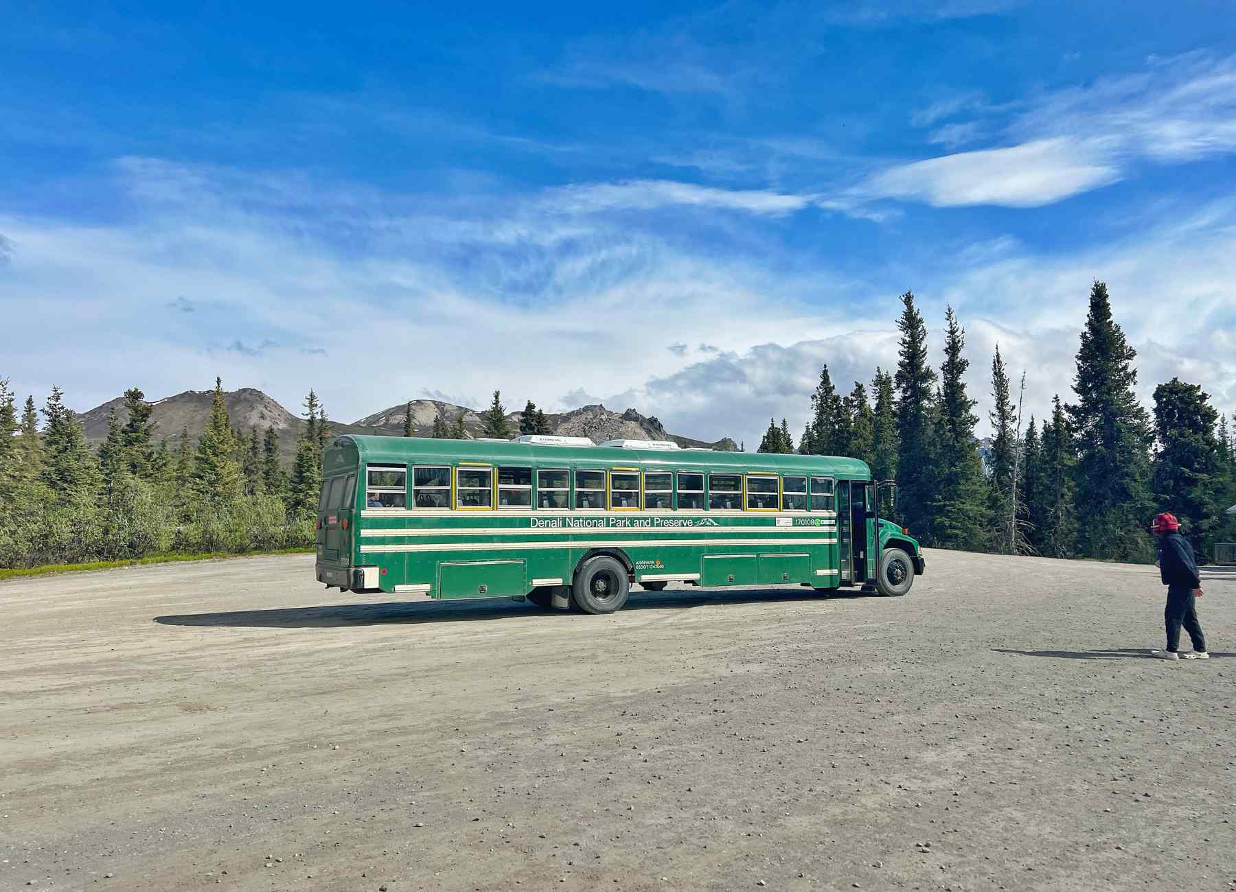 A Denali park shuttle stopped near the East Fork River turnaround with travelers stepping out to view wide tundra valleys and braided river channels under dramatic Alaska clouds, real photography