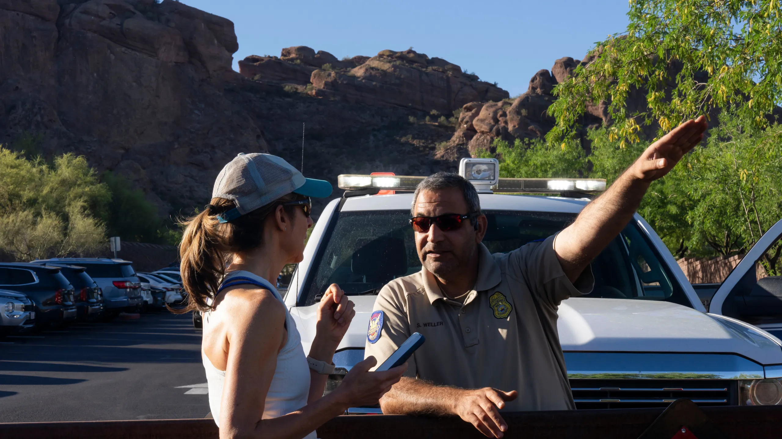 A National Park Service ranger speaking with a visitor holding a small quadcopter drone at a busy trailhead, mountains in the background, candid documentary photograph