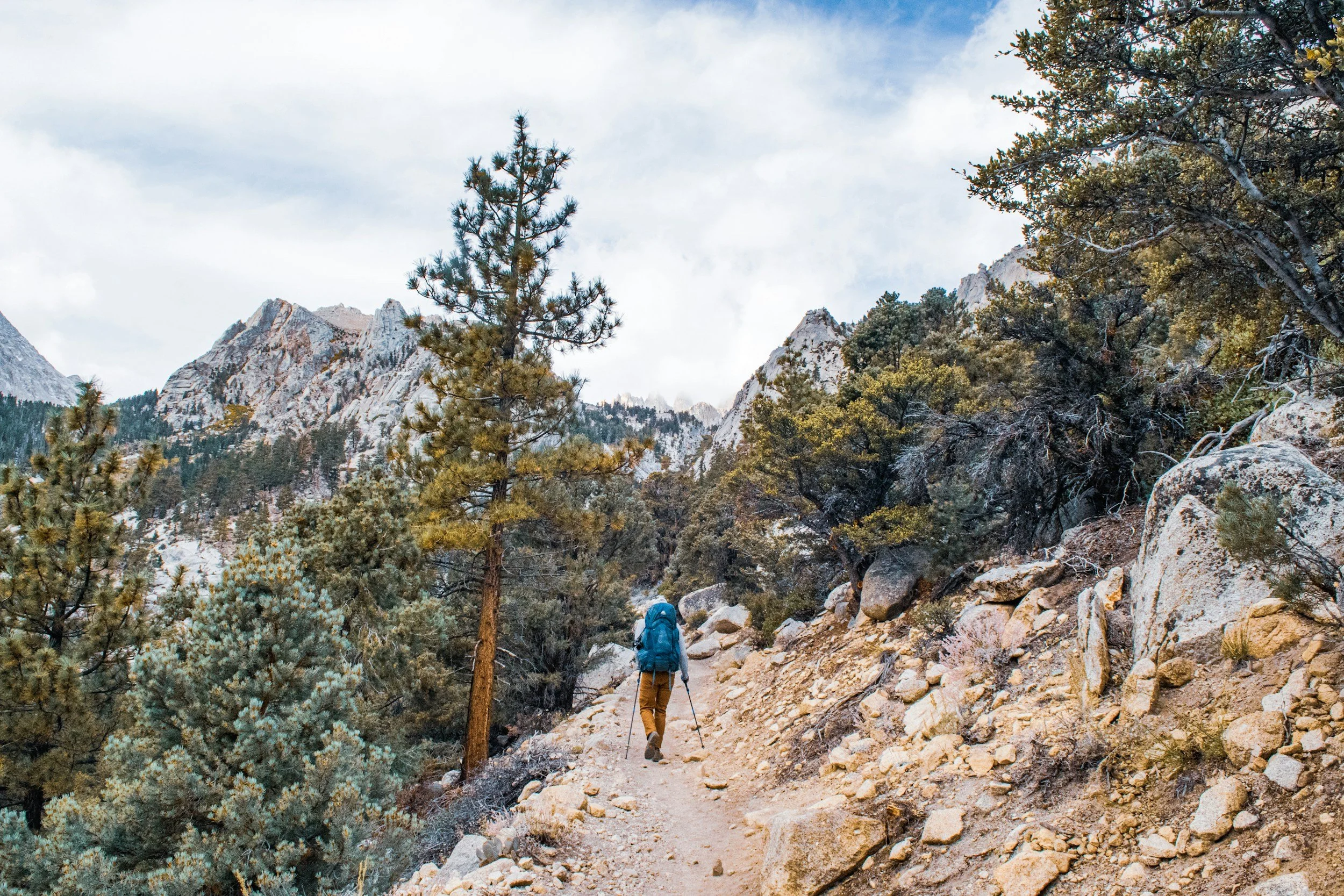 A backpacker stepping carefully across a rocky alpine trail with a full pack and trekking poles, rugged mountains in the distance under clear daylight, photorealistic