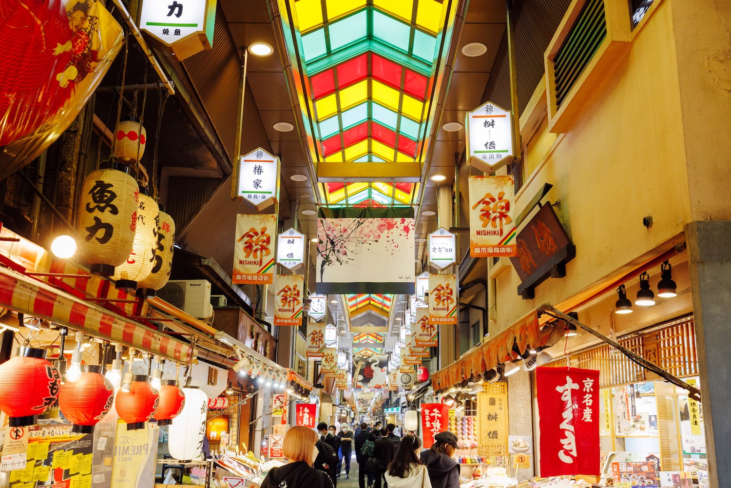 A bustling aisle inside Nishiki Market in Kyoto with hanging shop lights, food stalls on both sides, shoppers walking through, and colorful produce and snacks visible, photorealistic travel photography
