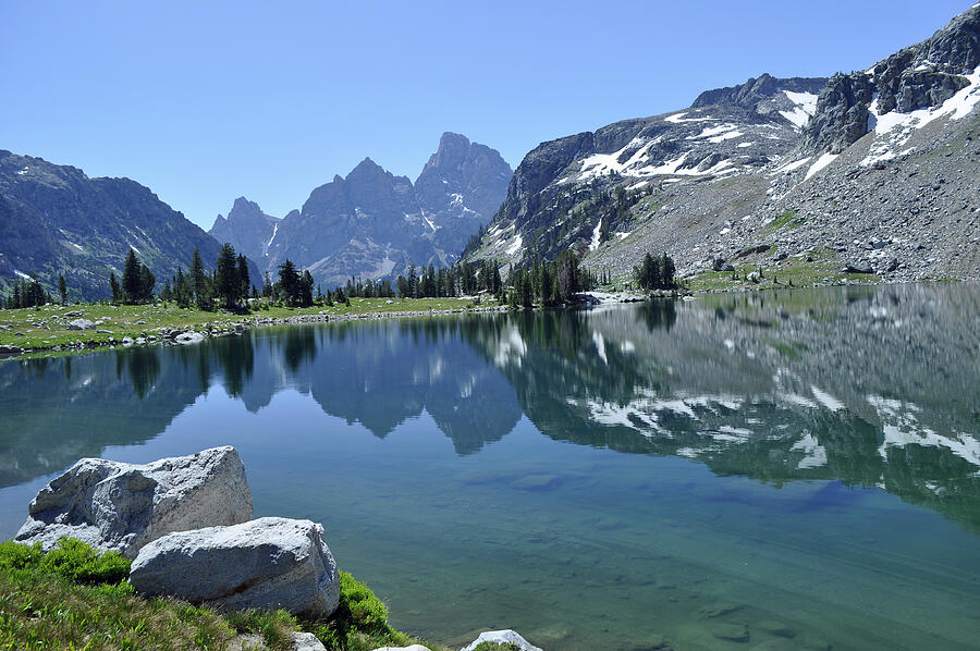 A calm Lake Solitude shoreline with smooth stones in the foreground and rugged Teton peaks reflected in the water under soft afternoon light, natural photo style