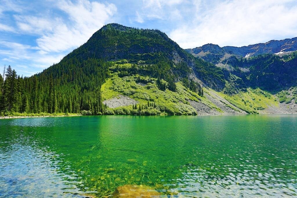 A calm mountain lake with evergreen forest and rugged peaks reflected in the water near the Rainy Lake trail in North Cascades National Park