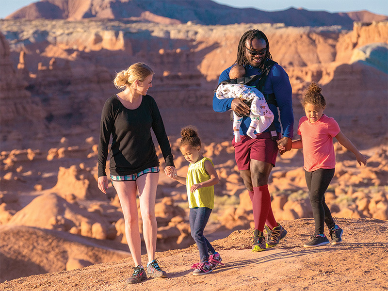 A candid travel photograph of a family walking between rounded sandstone hoodoos in the Valley of the Goblins, with kids in sun hats stepping from slickrock onto sandy ground