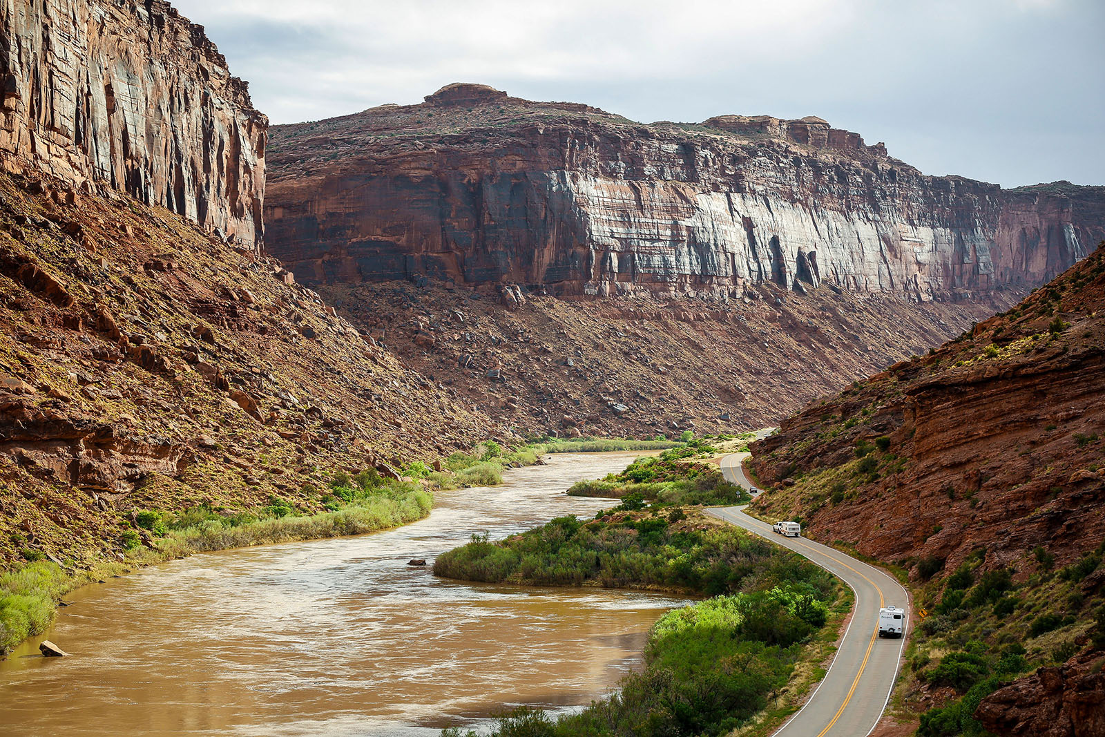 A car driving along Utah Highway 128 beside the Colorado River with red rock cliffs rising above the water in late morning light