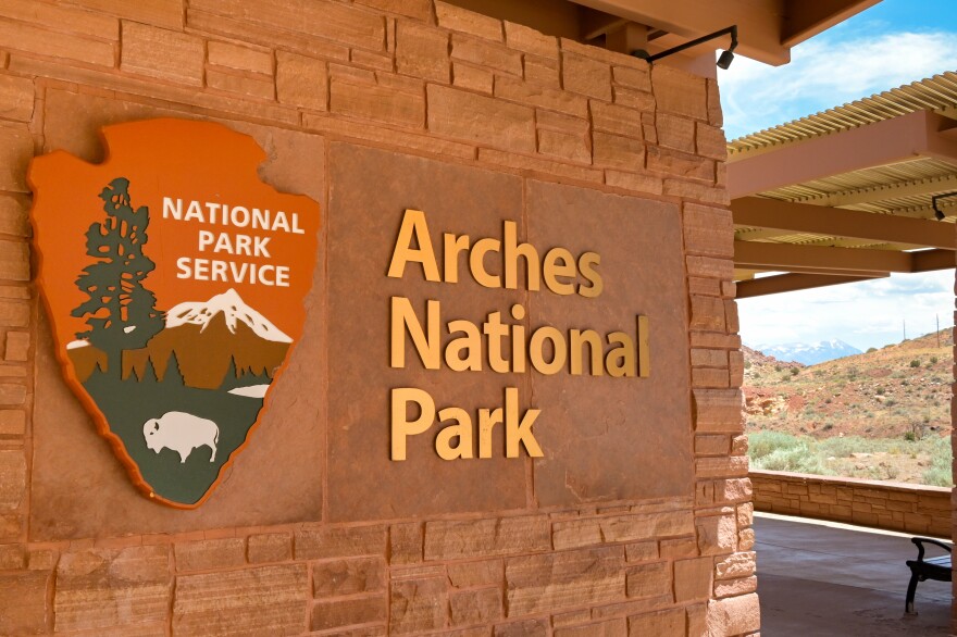 A car pulling up to the Arches National Park entrance station with red rock cliffs in the background, realistic travel photo