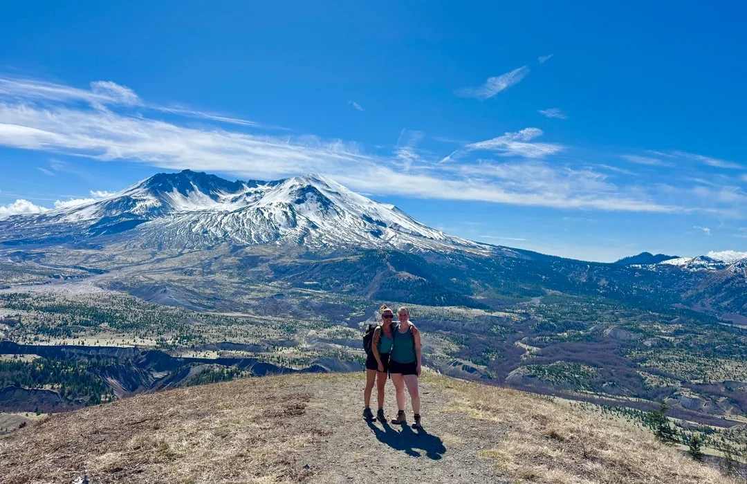A clear summer photograph of the Mount St. Helens crater viewed from the Johnston Ridge area, with the pumice plains in the foreground and hikers standing near a viewpoint railing