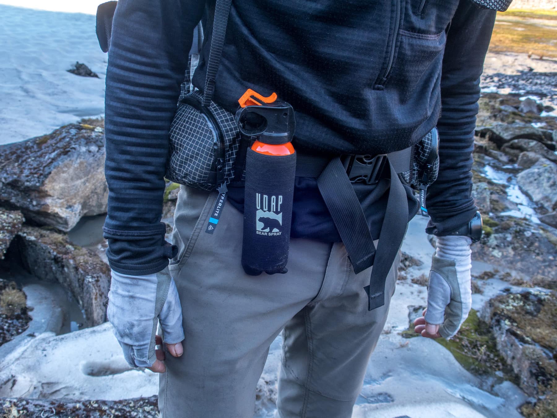 A close-up photo of a backpacker wearing a bear spray canister in a holster mounted on the hip belt of a hiking backpack, forest trail background