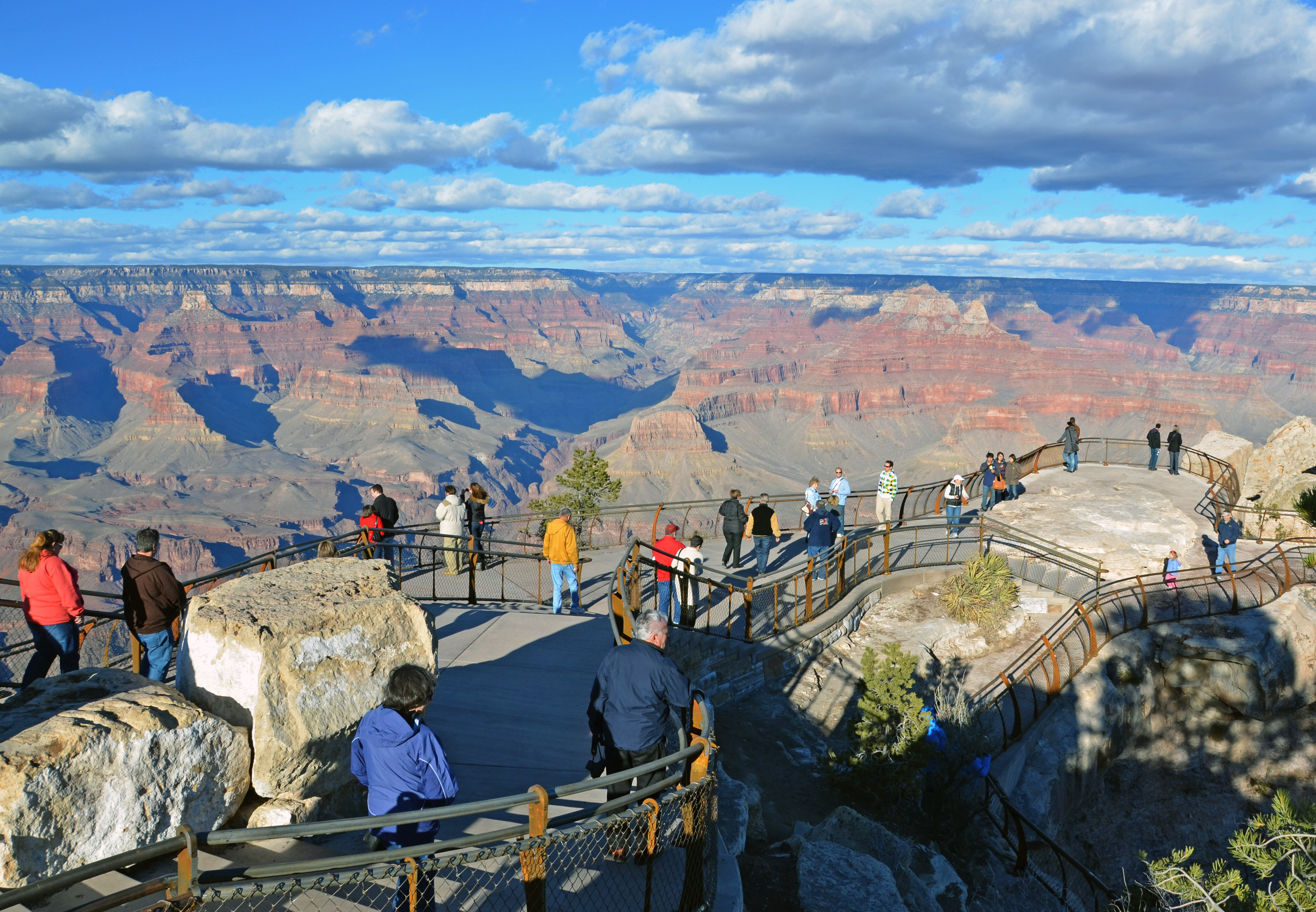 A crowd of visitors standing at Mather Point overlook railing looking into the Grand Canyon under clear skies, expansive layered rock formations, candid documentary travel photo