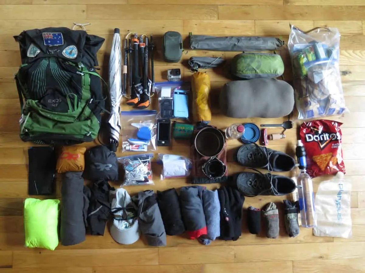 A day hiker at a trailhead placing a compact rain jacket, small first aid kit, and a collapsible water bottle into a small daypack beside trekking poles, realistic outdoor photography