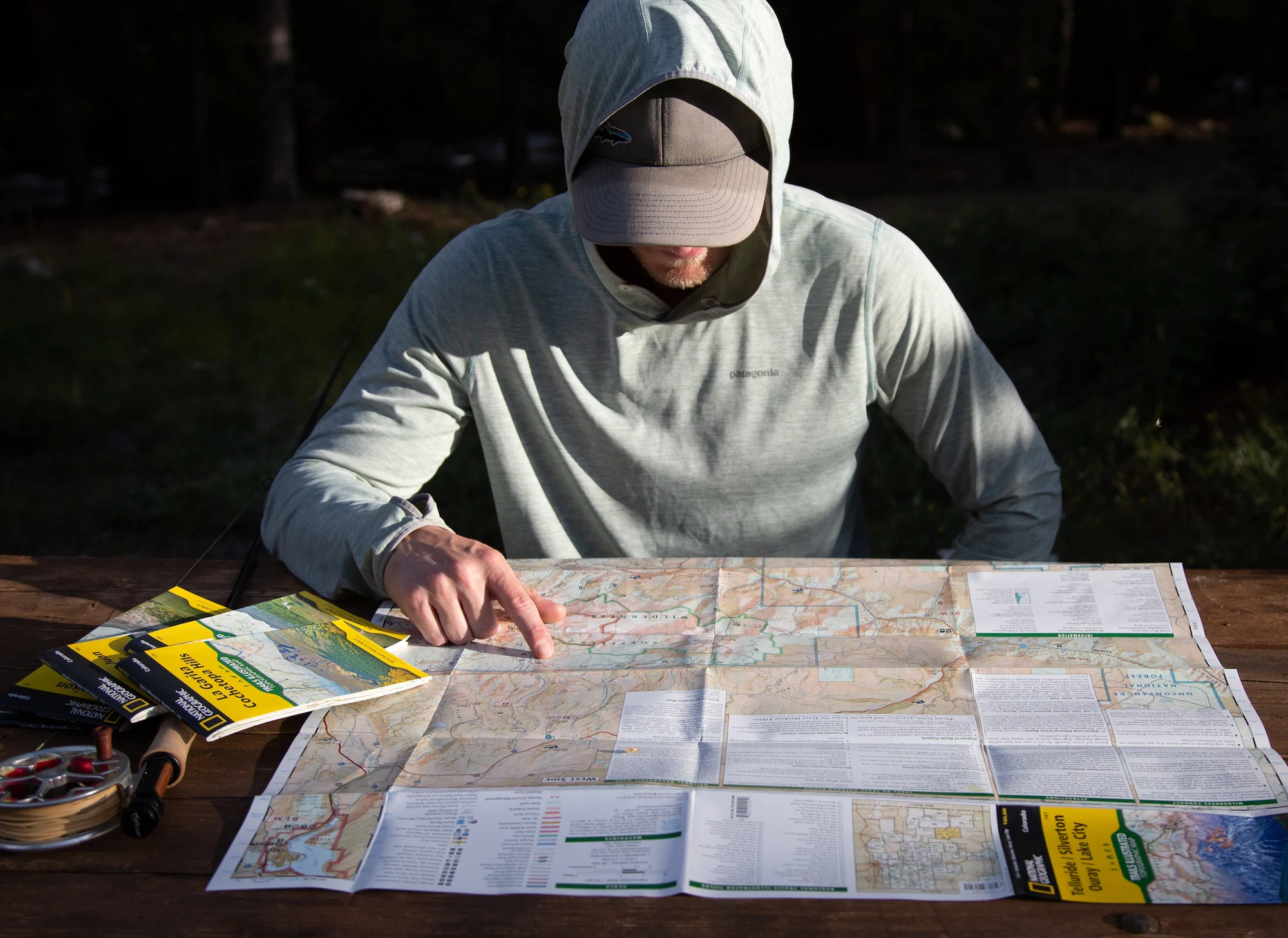 A day hiker standing at a trailhead holding a folded topographic map and a compass, with a forested hillside rising behind them in natural morning light, realistic photography