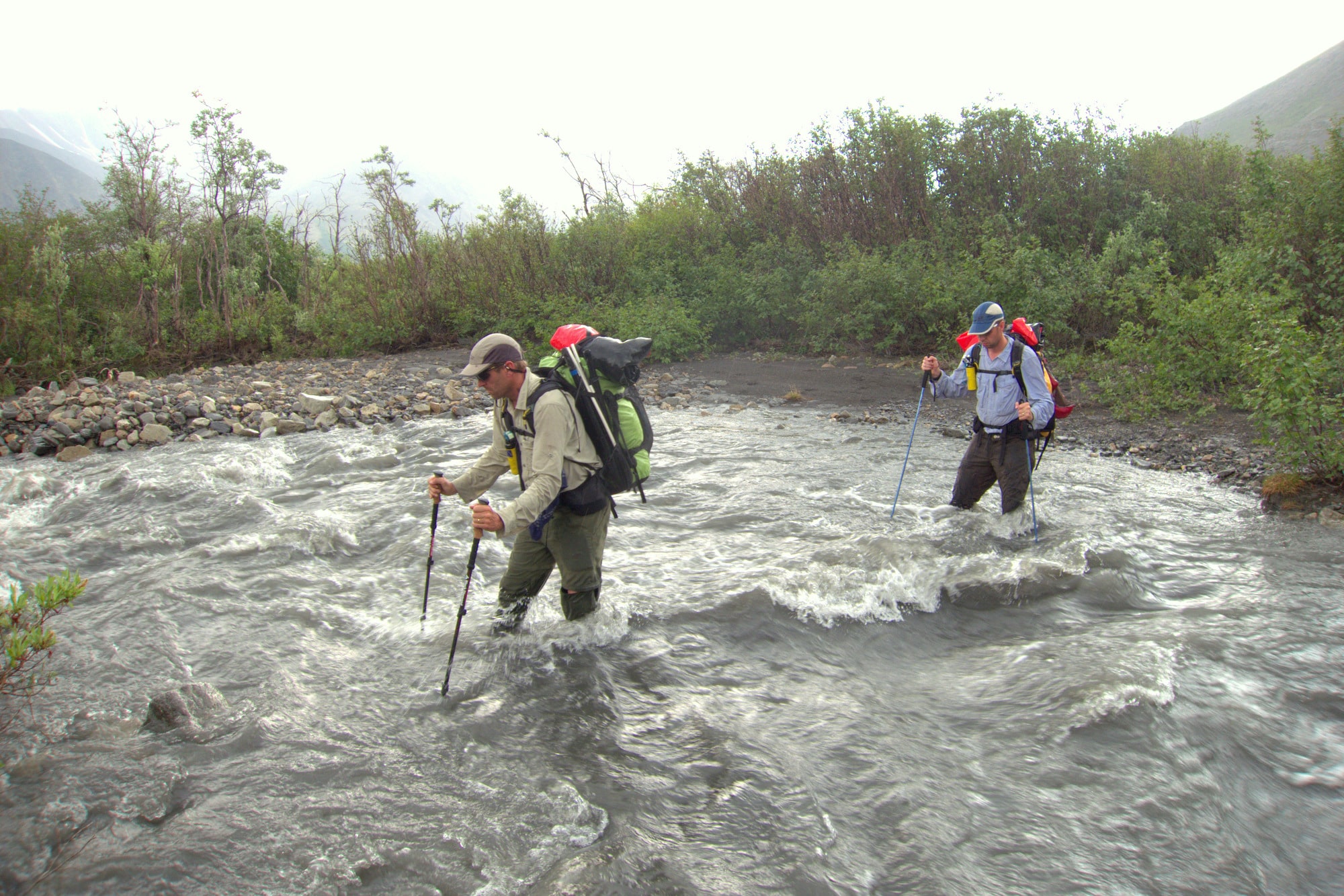 A day hiker standing on a rocky riverbank scouting a shallow stream crossing while holding trekking poles, with a visible current and forested mountains in the background, natural light photo