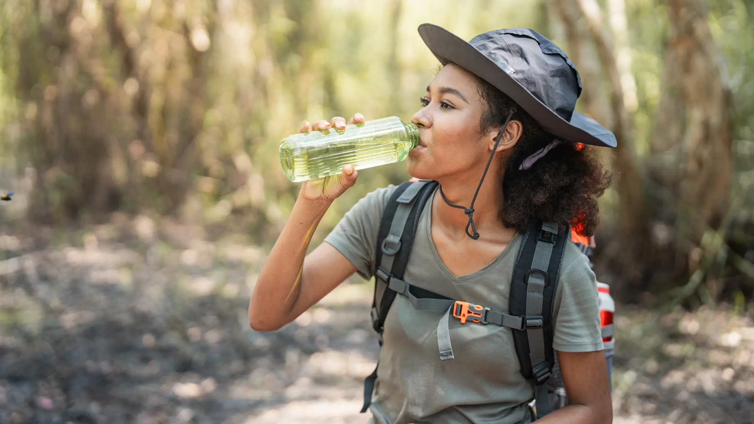 A day hiker wearing a small backpack and drinking from a hydration tube while walking on a dusty trail with midday sun, realistic outdoor photography style