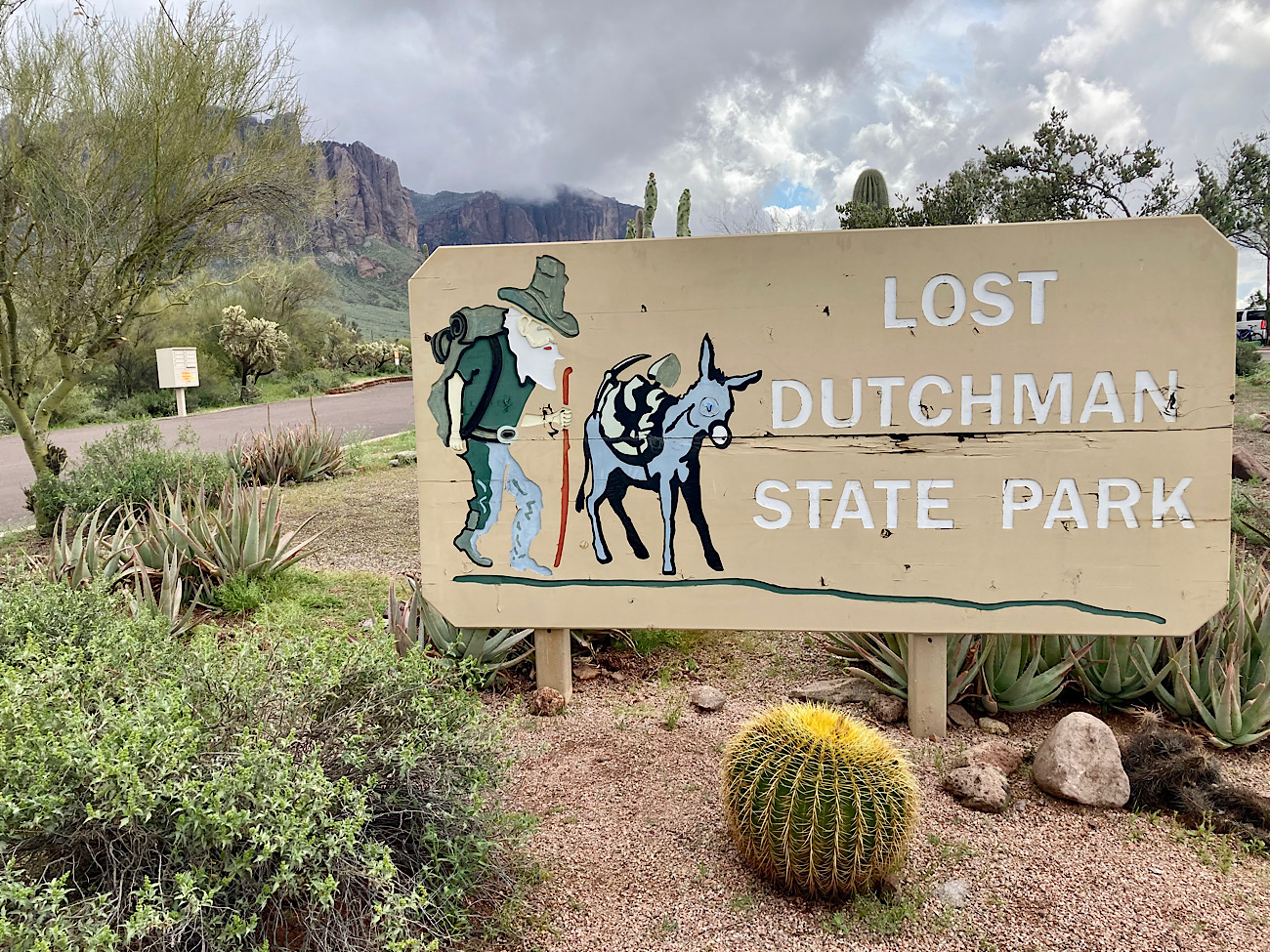 A daylight photograph of a Lost Dutchman State Park trailhead area in Apache Junction with a visible park sign and desert landscaping, with the Superstition Mountains in the distance