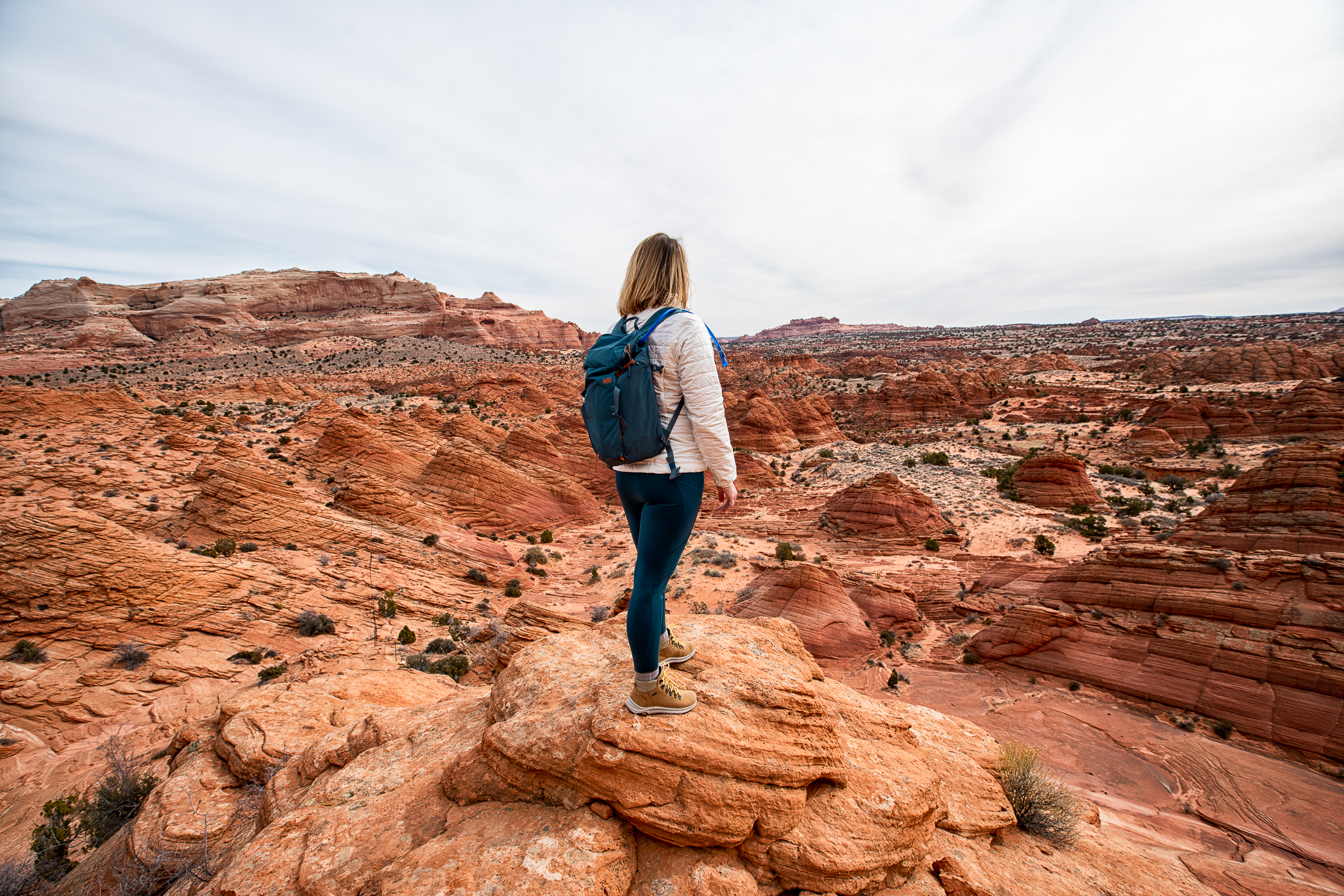 A desert hiker wearing a light sun hoodie and wide brim hat walking across sandy terrain with a full hydration pack, midday light in the Arizona Utah desert