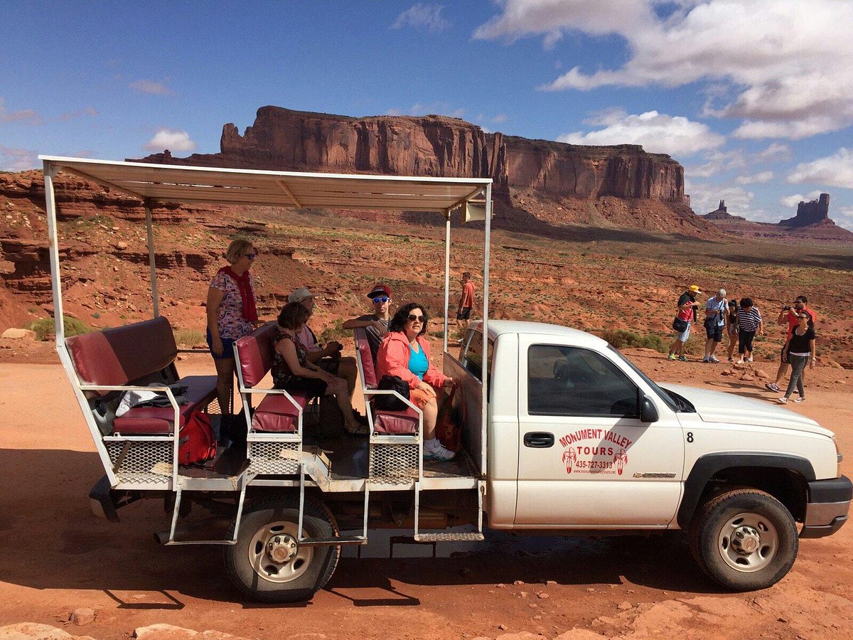 A documentary-style photograph of a Navajo guided tour vehicle driving along a sandy track in Monument Valley with towering buttes and warm late-afternoon light