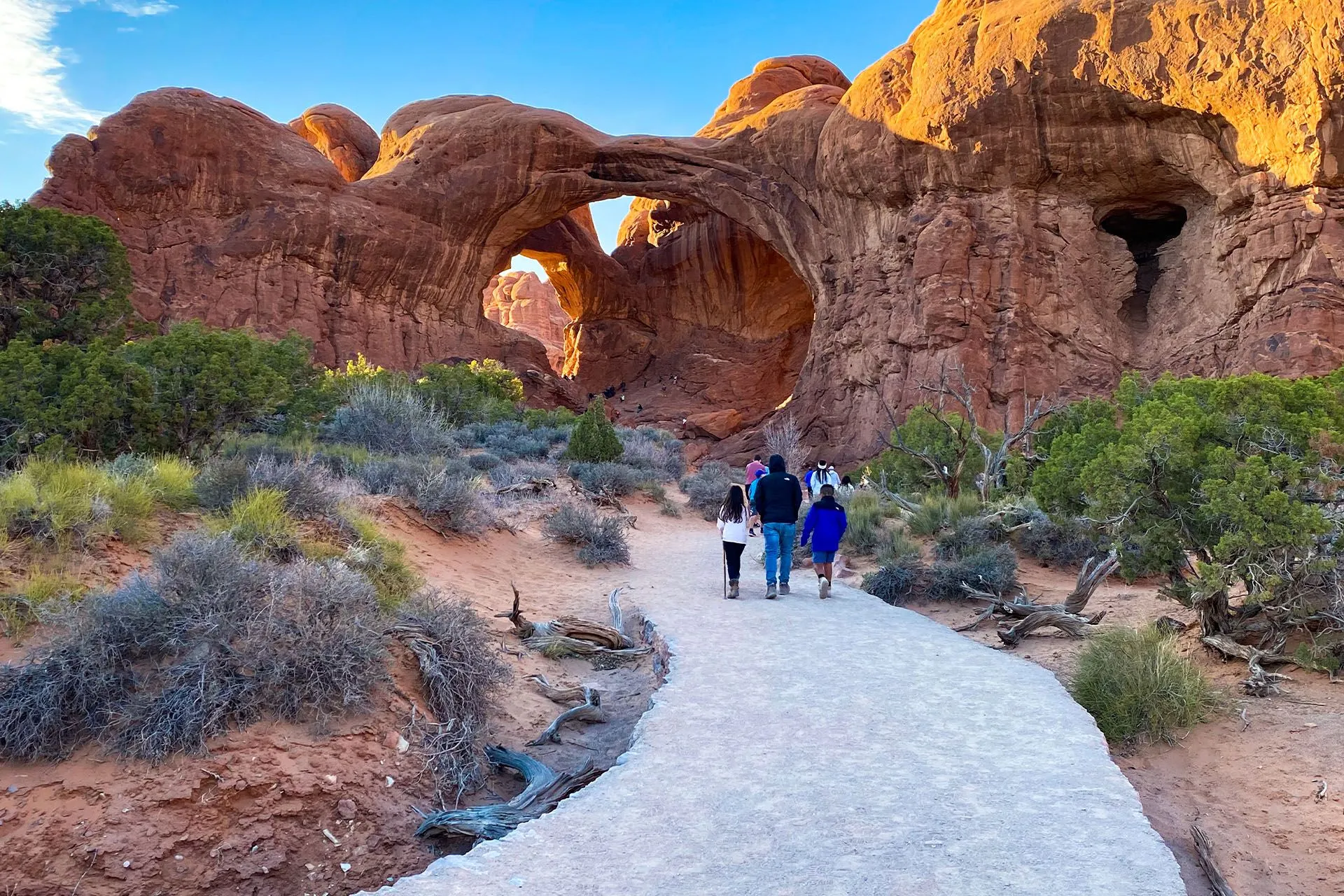 A family walking on a sandy trail toward Double Arch in Arches National Park under bright desert light, real travel photography style