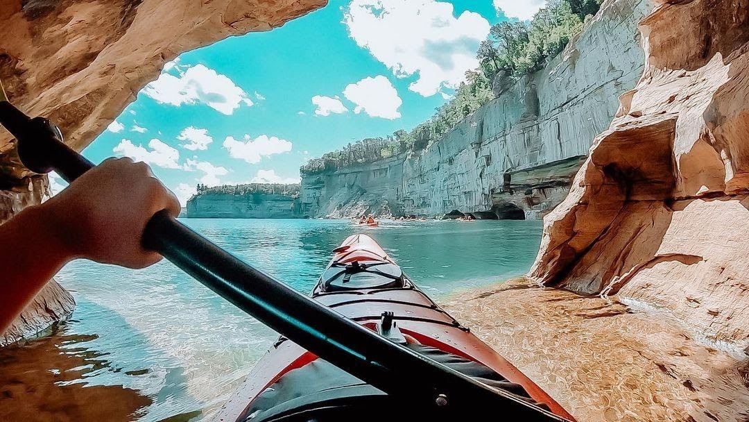 A guided sea kayak group paddling close to the colorful Pictured Rocks cliffs on a calm day, with turquoise water and sandstone streaks visible behind them