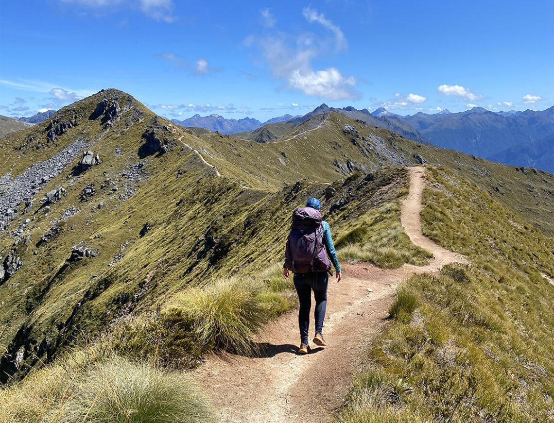 A hiker at a sunny summer trailhead wearing a lightweight long-sleeve sun shirt, breathable hiking pants, a wide-brim hat, sunglasses, and a small daypack, real photography style