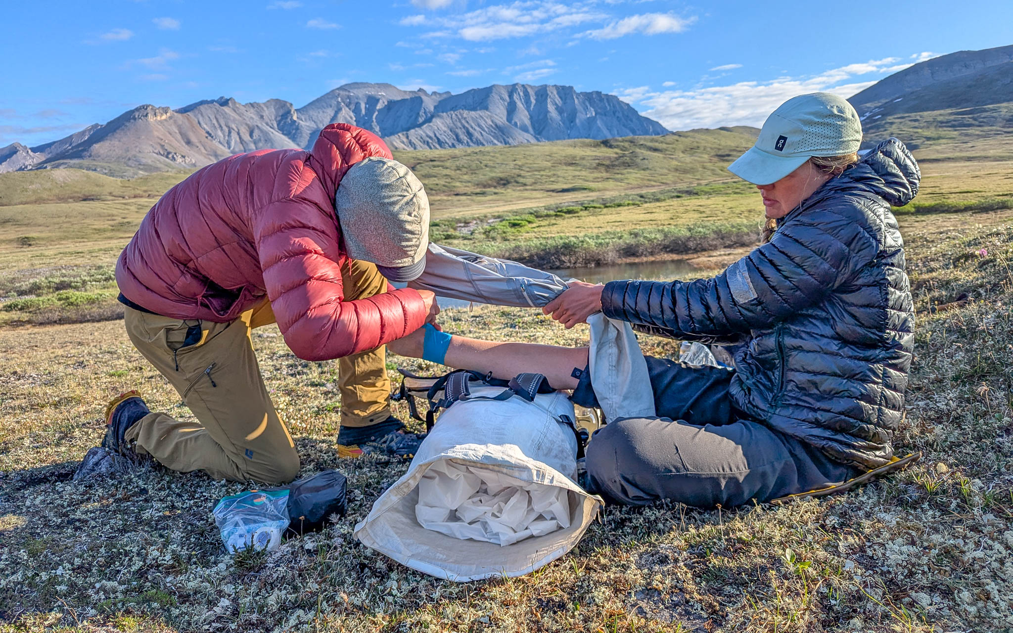 A hiker kneeling beside a seated companion on a mountain trail, placing a wet bandana on the companion's neck while another person holds an open water bottle, realistic outdoor photography
