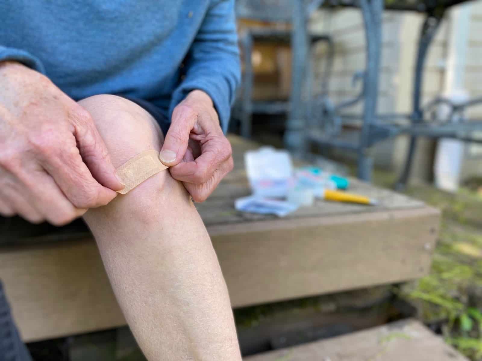 A hiker kneeling on a forest trail rinsing a scraped knee with water from a sports-cap bottle while another person holds gauze nearby, natural candid photograph