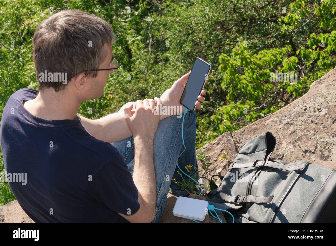 A hiker sitting on a flat rock beside a mountain trail charging a smartphone from a compact power bank, with a backpack open and alpine scenery in the background, realistic outdoor photography