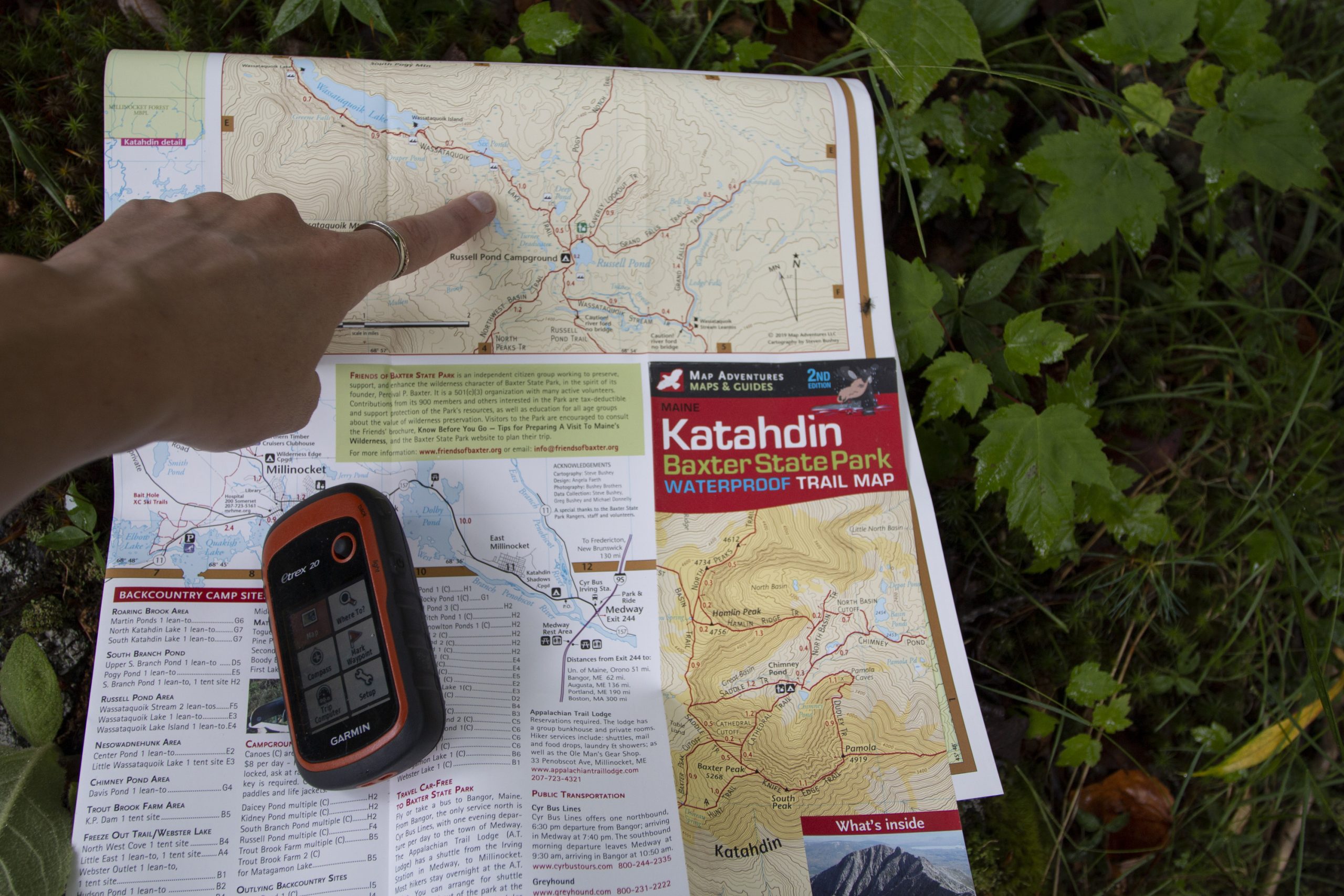 A hiker sitting on a log beside a mountain trail, holding a folded topographic map and comparing it to the surrounding ridgelines, realistic photography style