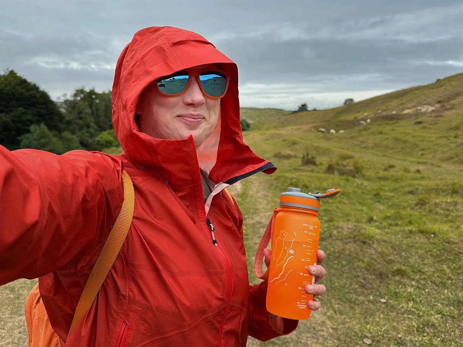 A hiker sitting on a sunlit rock ledge during a summer day hike, holding a reusable bottle with a sports cap and resting beside a backpack, realistic outdoor photography style