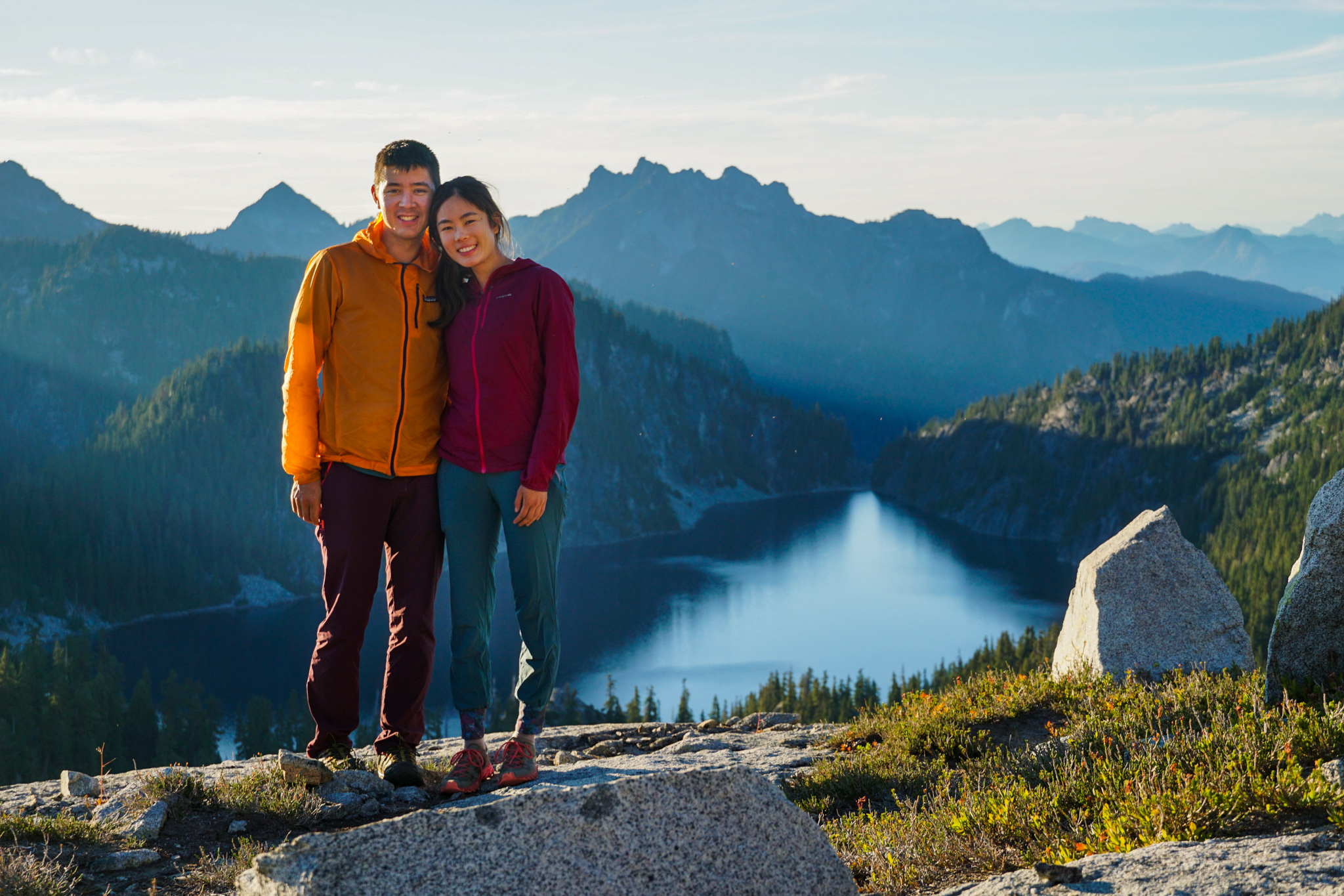 A hiker sitting on a sunny alpine trail taking a water break, with a daypack and trekking poles beside them and mountains in the background, real outdoor photography style