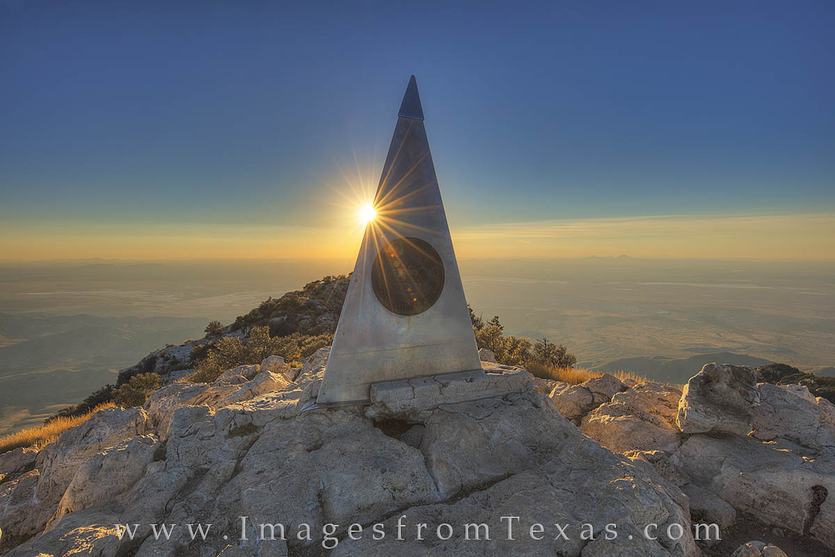 A hiker standing near the Guadalupe Peak summit marker on a bright day with expansive desert and mountain views stretching to the horizon