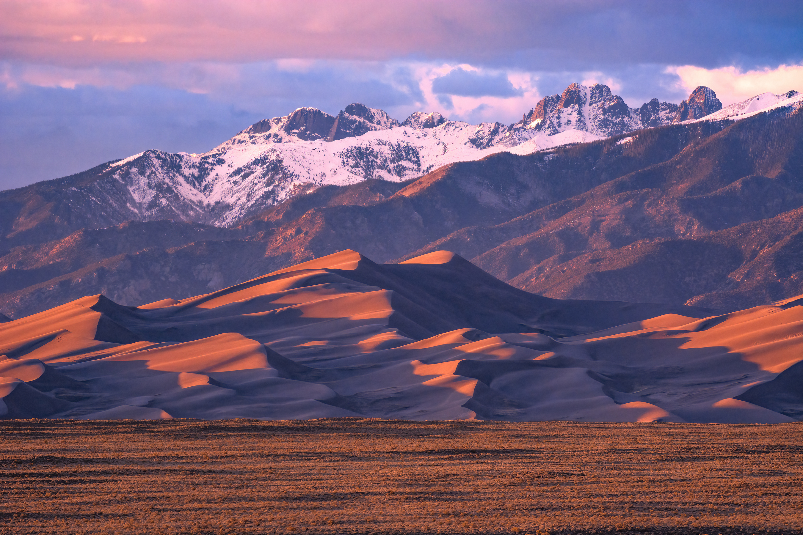 A hiker standing on the crest of High Dune at Great Sand Dunes National Park during sunset with rippled sand patterns in the foreground and the Sangre de Cristo Mountains in the distance