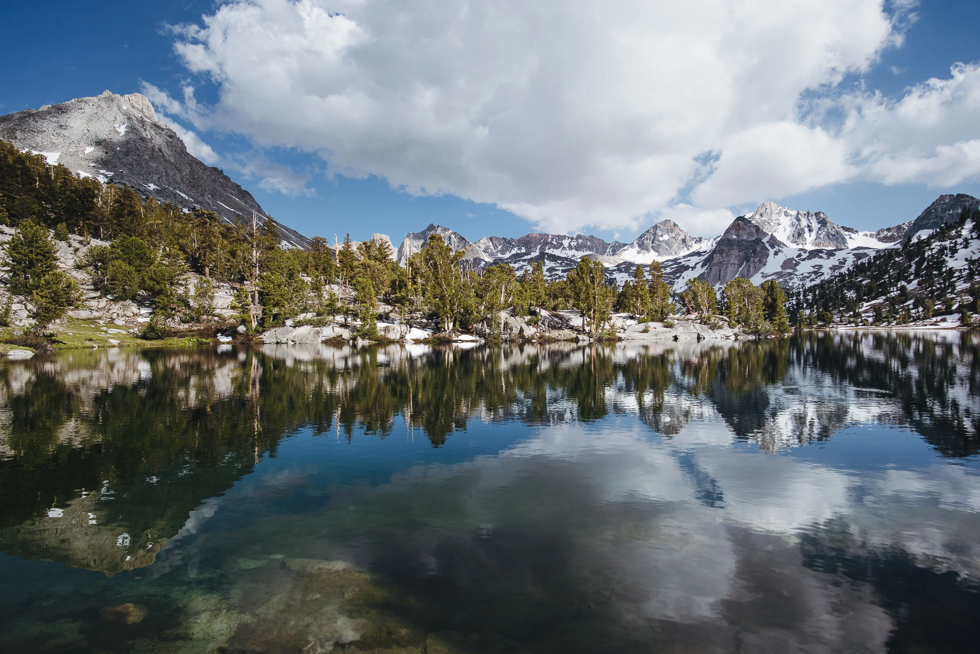 A hiker walking beside a clear mountain river on the Rae Lakes Loop trail near Road's End in Kings Canyon National Park with pine forest and granite slopes, realistic outdoor photograph