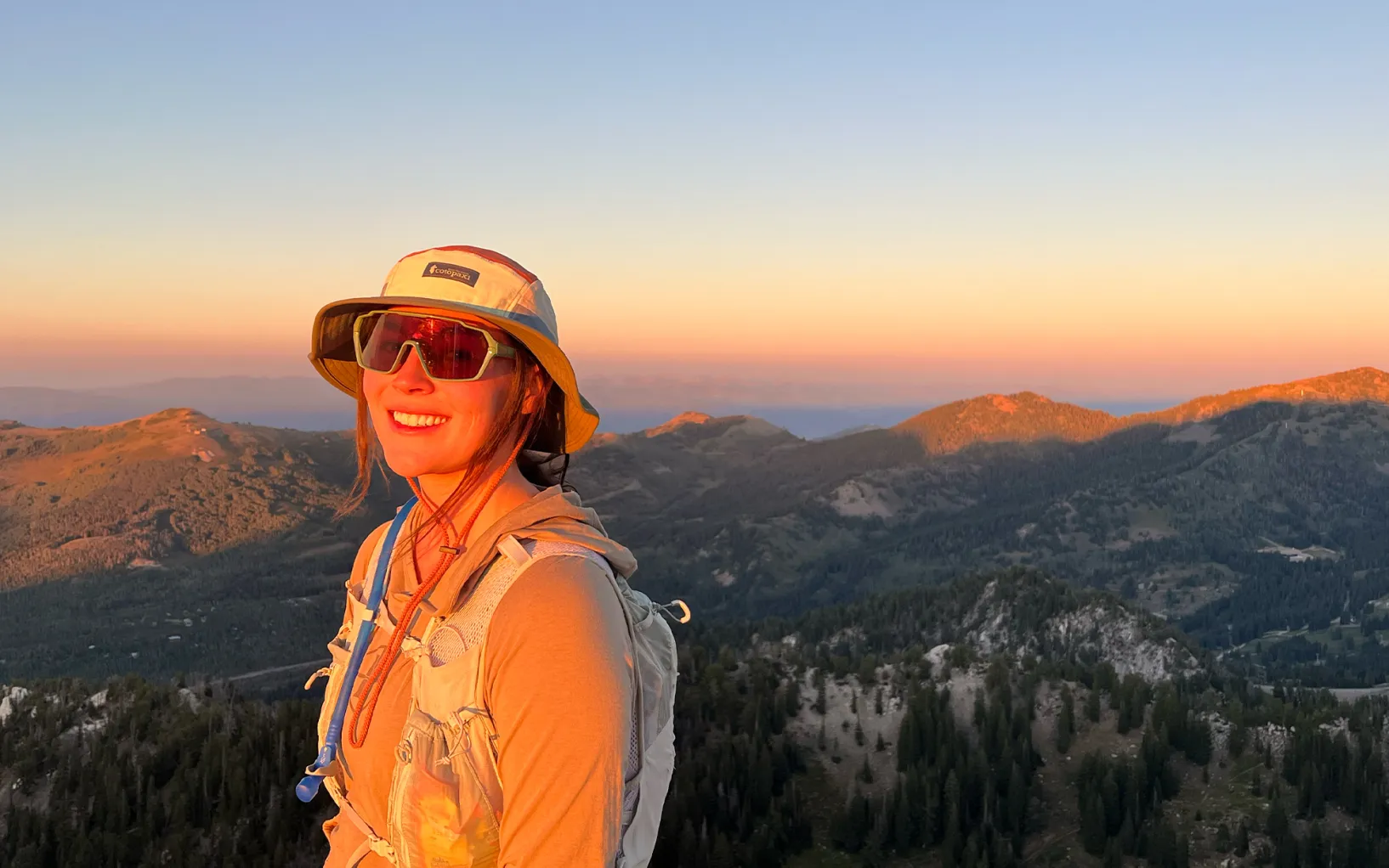 A hiker wearing a wide-brim hat and long-sleeve sun shirt applying sunscreen to their face beside a desert trailhead sign in bright morning light