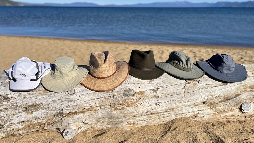 A hiker wearing a wide-brim sun hat and light long-sleeve shirt walking on a dry canyon trail with strong sunlight and sparse shade, realistic outdoor photography