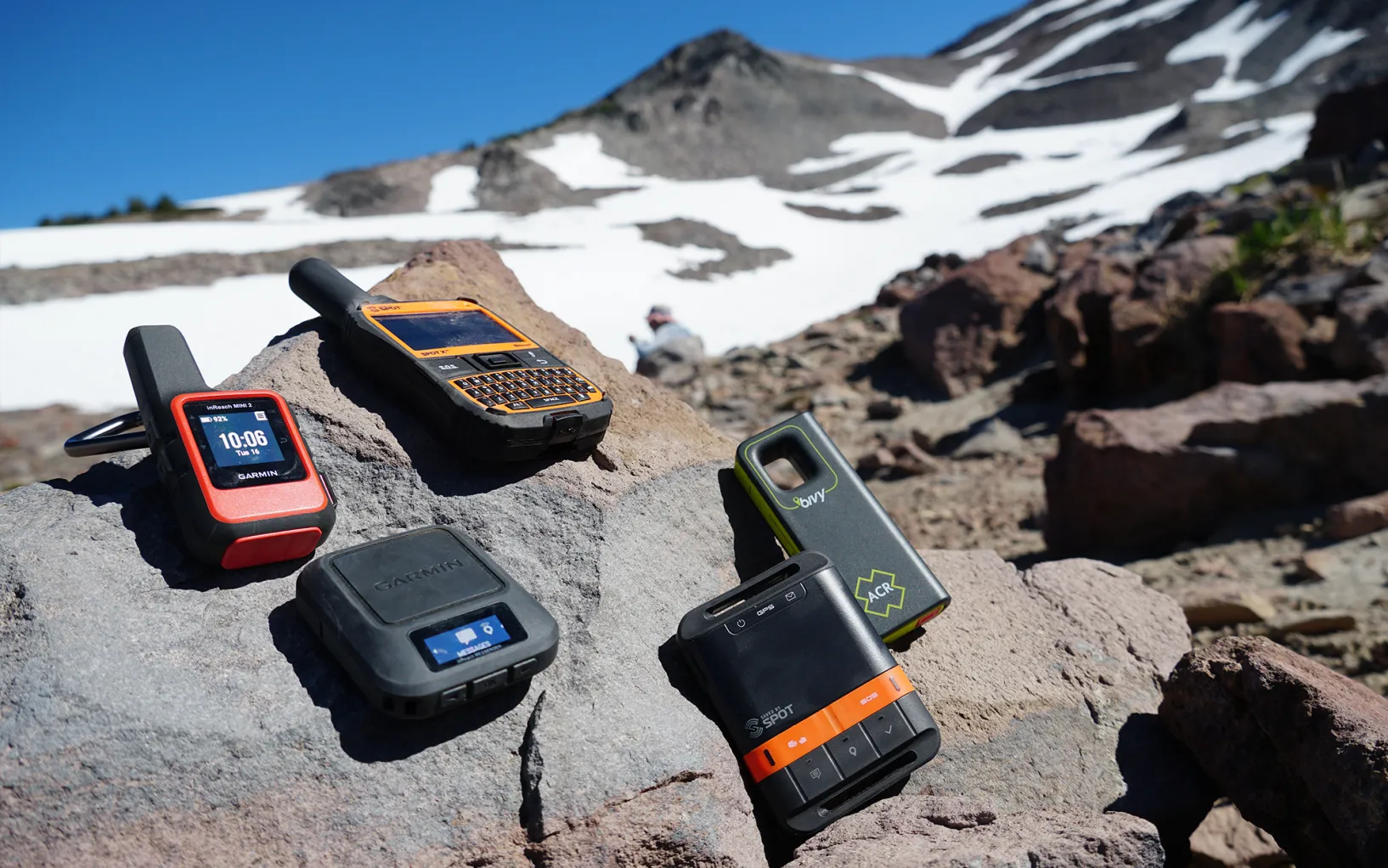 A hiker wearing a wind jacket holding a handheld satellite messenger device while standing on a windy mountain ridge under a cloudy sky, realistic outdoor photography
