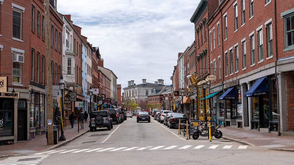 A lively but uncrowded downtown street in Portsmouth, New Hampshire with brick storefronts, fall decorations, and pedestrians in light jackets under overcast coastal light, photorealistic travel photography