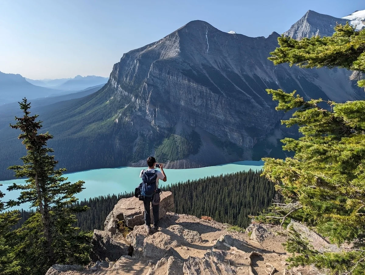 A lone hiker stands on a rocky viewpoint above layered mountain ridgelines and a winding river valley in crisp afternoon light.