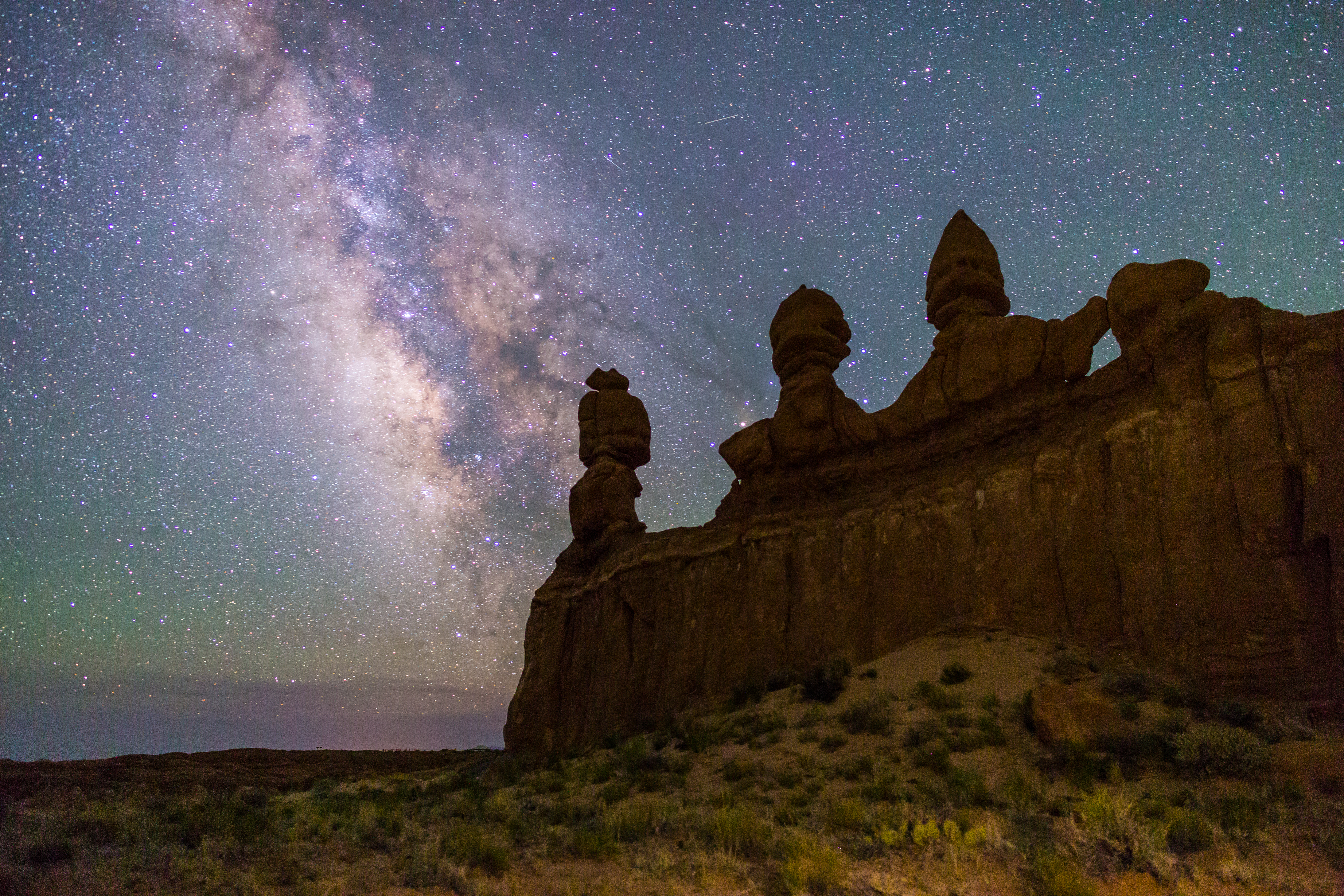 A long-exposure style night photograph from Goblin Valley showing silhouetted hoodoos under a star-filled sky, with a person sitting quietly on a blanket in the foreground