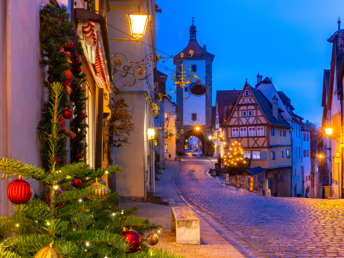A narrow cobblestone street in Rothenburg ob der Tauber at night, lined with half-timbered houses decorated with evergreen garlands and warm Christmas lights, with a light dusting of snow, photorealistic travel photography