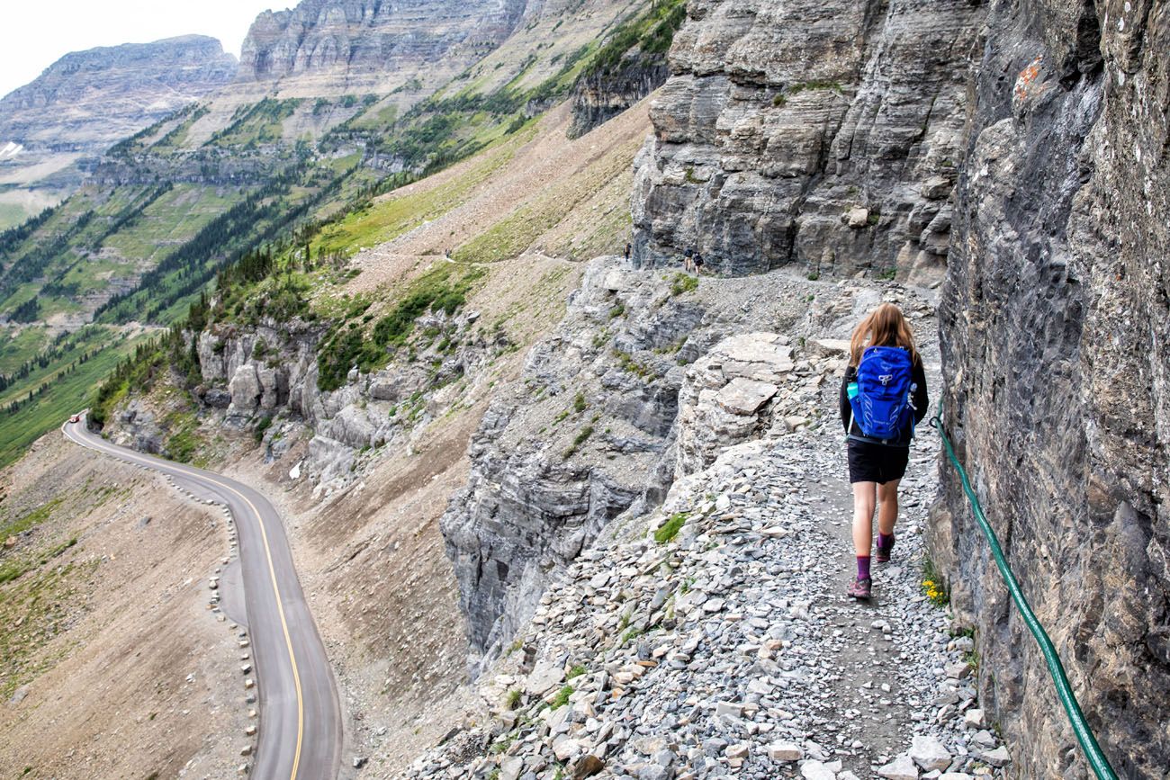 A narrow rocky trail on the Highline Trail near Logan Pass with a steel safety cable bolted into the cliff, steep mountain drop to one side, realistic outdoor photo