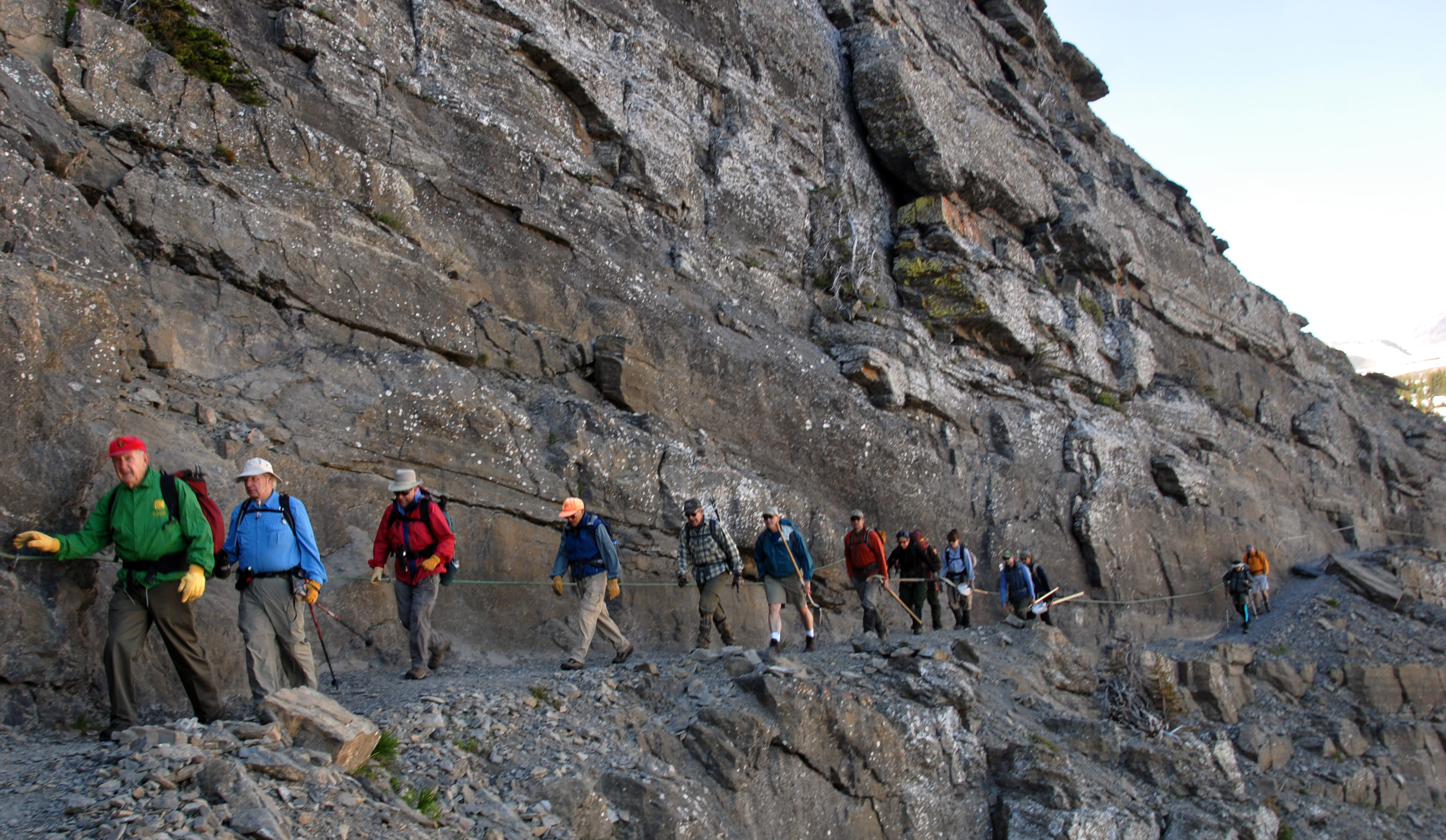 A narrow section of the Highline Trail cut into a steep mountainside with a hand cable along the rock wall, hikers spaced along the trail, and a deep valley view under clear summer light, photorealistic