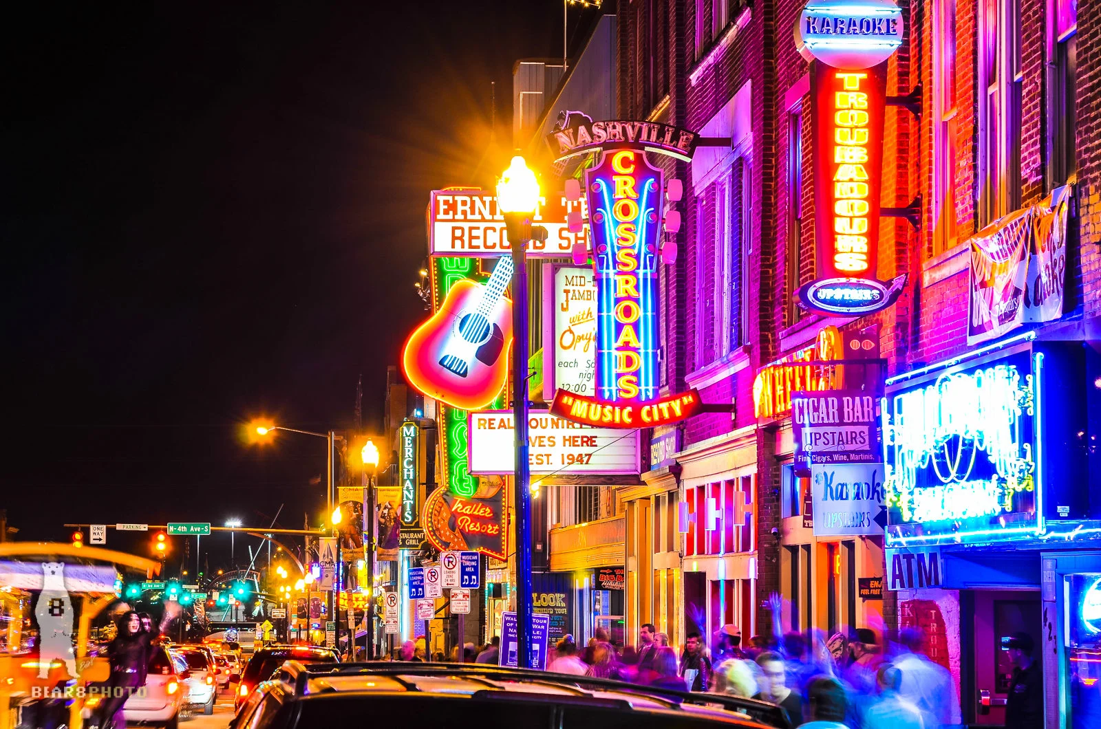 A nighttime street scene on Broadway in Nashville with glowing neon honky-tonk signs, live music spilling onto the sidewalk, and pedestrians walking past bars