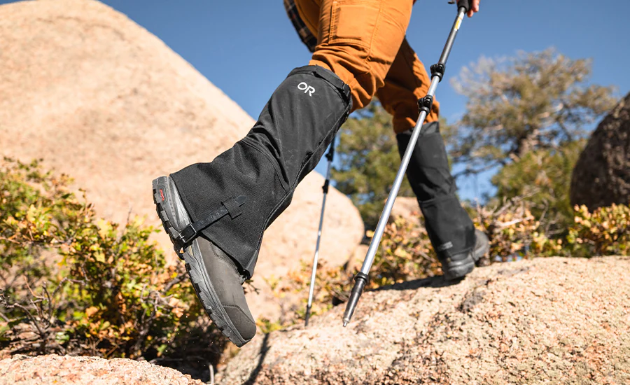 A pair of hiking boots with protective gaiters beside a backpack at a forest trail rest stop, leaf litter and rocks visible, real outdoor photography style