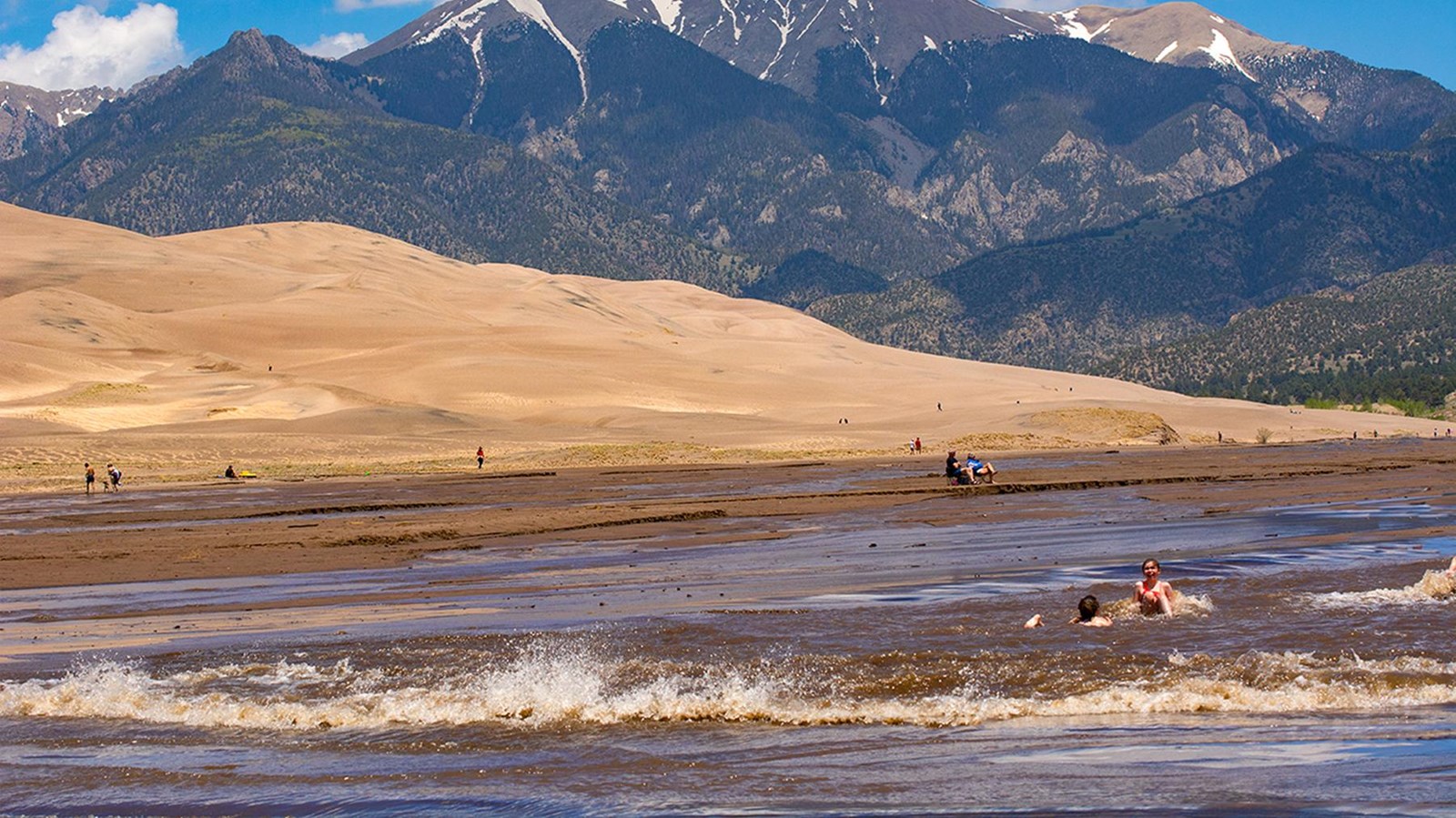 A parent and child crouched beside Medano Creek building a small sand dam with shallow water flowing around it at the base of the Great Sand Dunes, candid travel photography