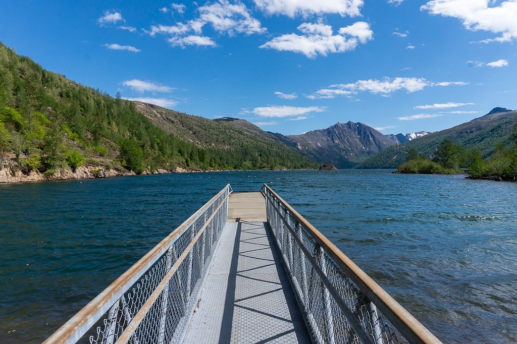 A photograph of Coldwater Lake on a clear day with calm water reflecting forested slopes and distant views toward the Mount St. Helens blast zone