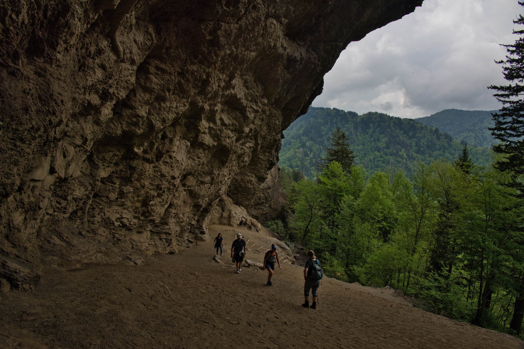 A photograph of hikers walking beneath the Alum Cave Bluffs overhang, with textured rock walls curving overhead and filtered sunlight illuminating the trail