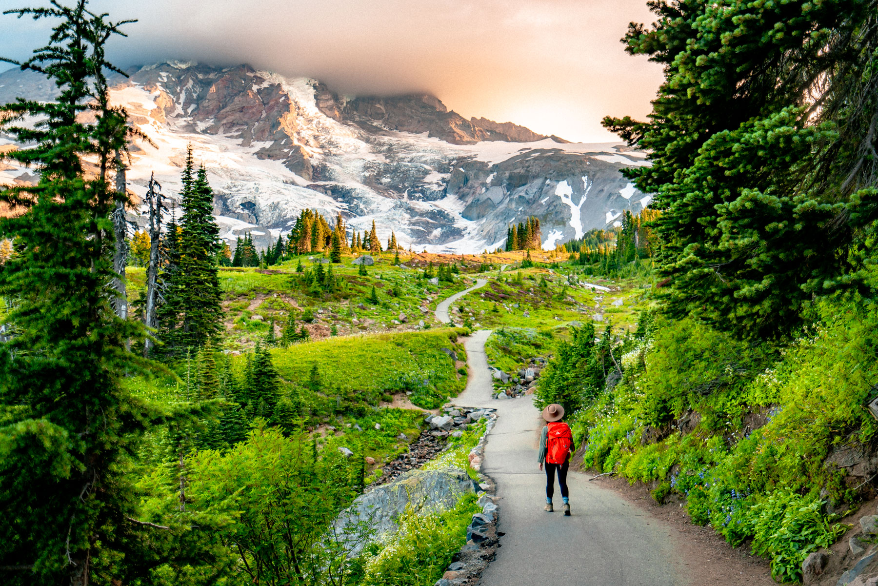 A photorealistic hiking trail in Mount Rainier National Park with wildflowers along the path and Mount Rainier towering in the background under a partly cloudy sky, travel photography style