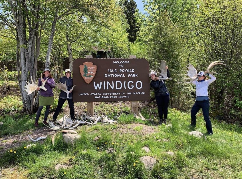 A real-life photograph of hikers stepping off a ferry at the Windigo dock on Isle Royale with backpacks, calm water, and dense green forest in the background