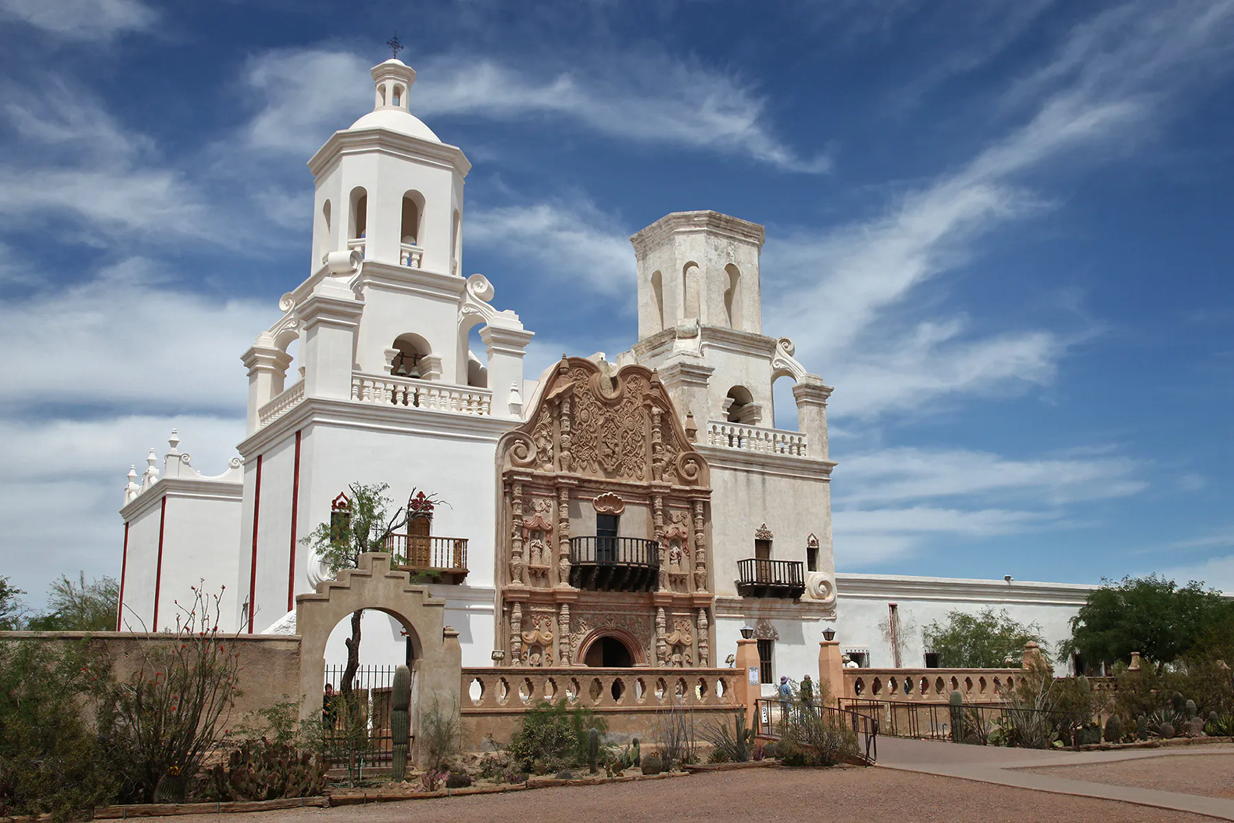 A real-life photograph of the white San Xavier del Bac Mission facade glowing in afternoon sun with desert vegetation in the foreground and a deep blue Arizona sky
