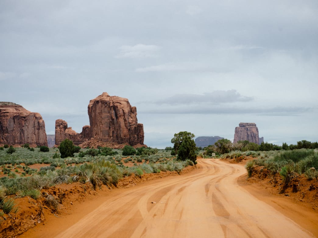 A real-life travel photograph of the unpaved scenic drive road in Monument Valley Tribal Park, with a vehicle kicking up dust and tall red buttes in the distance