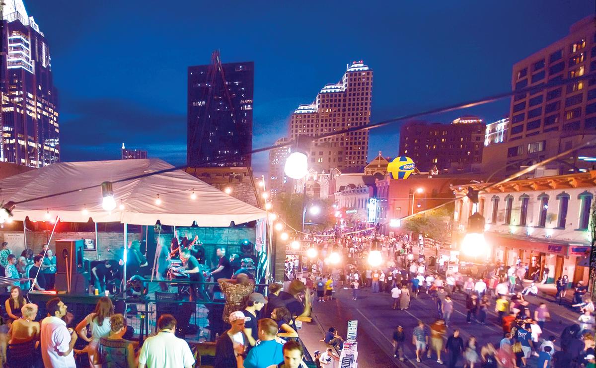 A real nighttime photo on Austin’s Sixth Street with neon-lit bar fronts, pedestrians on the sidewalk, and a live music venue entrance glowing in the background